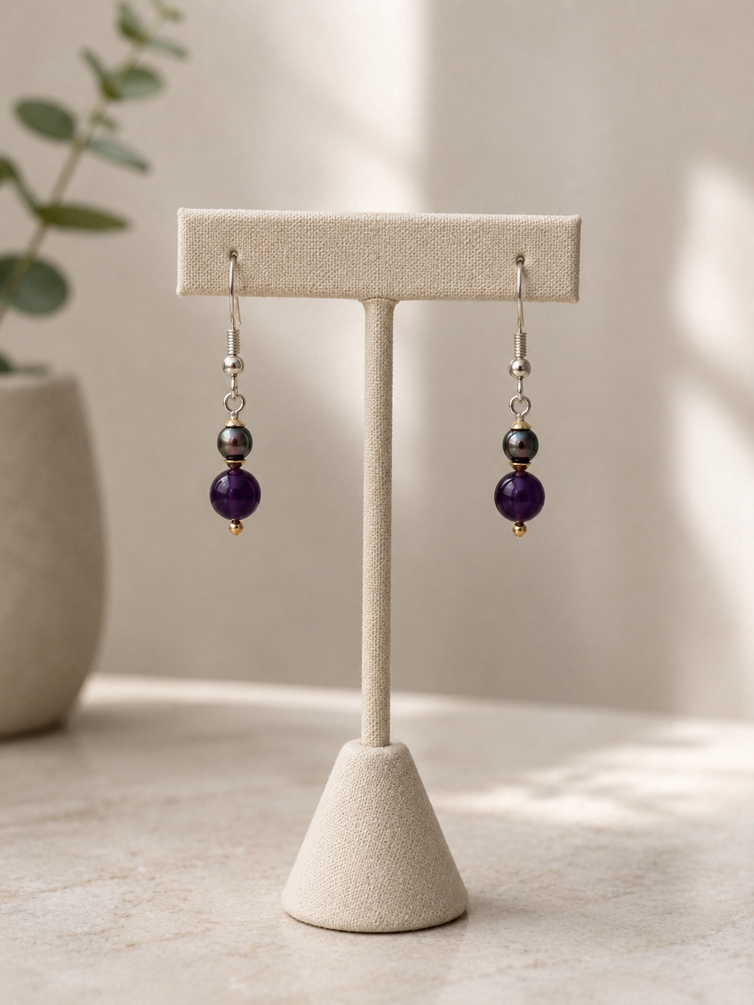 Handcrafted gemstone drop earrings displayed on a textured cream-colored jewelry stand against a soft, naturally lit background. Each earring features a deep purple amethyst bead paired with a dark iridescent pearl, accented by delicate gold-tone details and suspended from 925 sterling silver hooks. The elegant combination of rich gemstone hues and lustrous pearl tones creates a refined and timeless design. Graceful and versatile, these earrings are perfect for special occasions or adding a touch of sophist