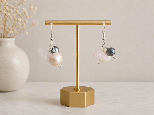 Handcrafted drop earrings displayed on a gold jewelry stand against a soft neutral background. Each earring features a luminous white coin pearl paired with a dark gray pearl and a pale pink rose quartz nugget, suspended from 925 sterling silver hooks. The elegant combination of pearls and natural gemstones creates a delicate, romantic design.