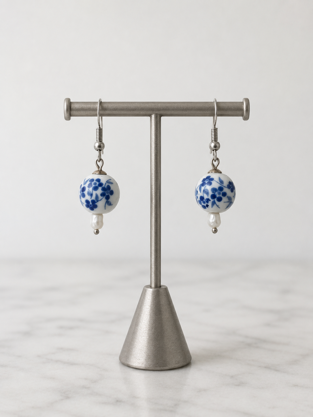 Handcrafted ceramic and pearl drop earrings displayed on a brushed silver jewelry stand against a soft neutral background. Each earring features a round white ceramic bead adorned with delicate blue floral motifs, paired with a luminous freshwater pearl drop and suspended from 925 sterling silver hooks. Inspired by classic porcelain artistry, the earrings blend timeless elegance with handcrafted charm. Their delicate blue-and-white design evokes sophistication and serenity, making them a graceful accessory 